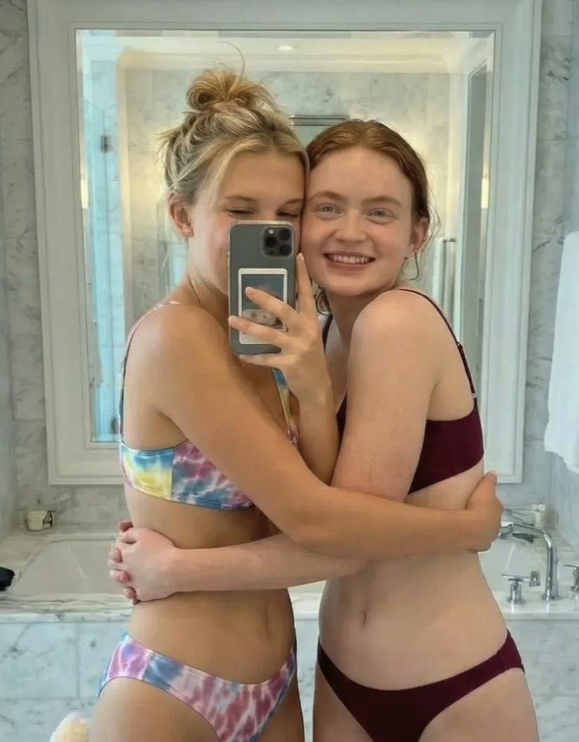 Two young women resembling Millie Bobby Brown and Sadie Sink embracing while taking a mirror selfie in a bathroom.