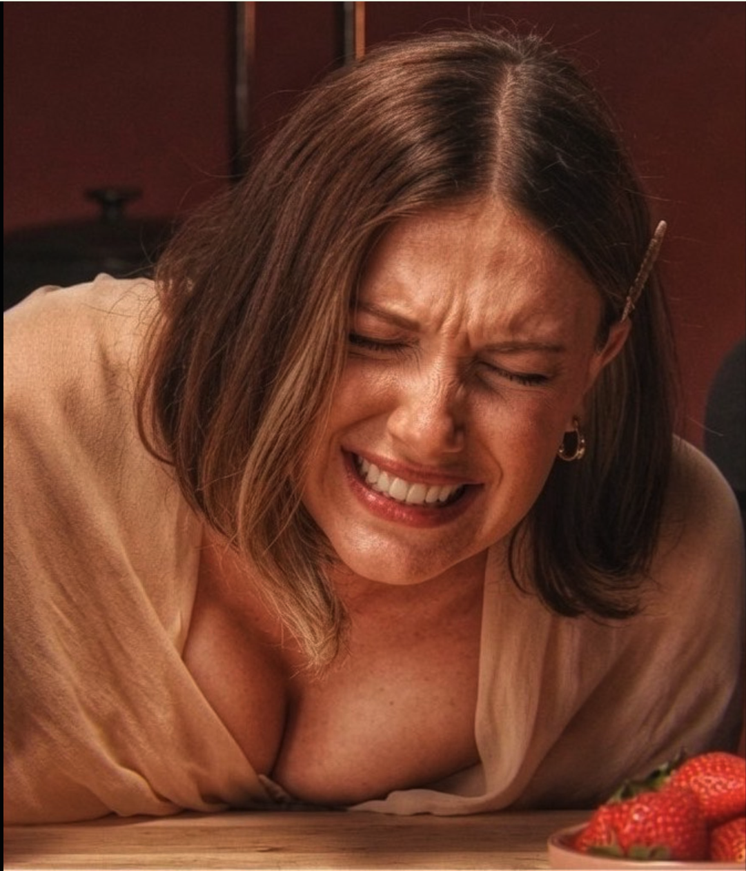 A woman resembling Millie Bobby Brown with eyes closed and mouth open in an exaggerated expression near a bowl of strawberries.