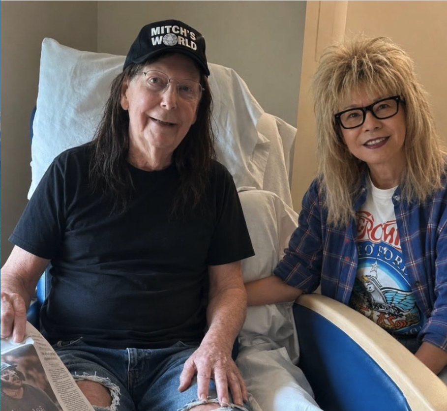 A man resembling Mitch McConnell wearing a 'Mitch's World' cap seated in a hospital bed beside a woman resembling Elaine Chao.