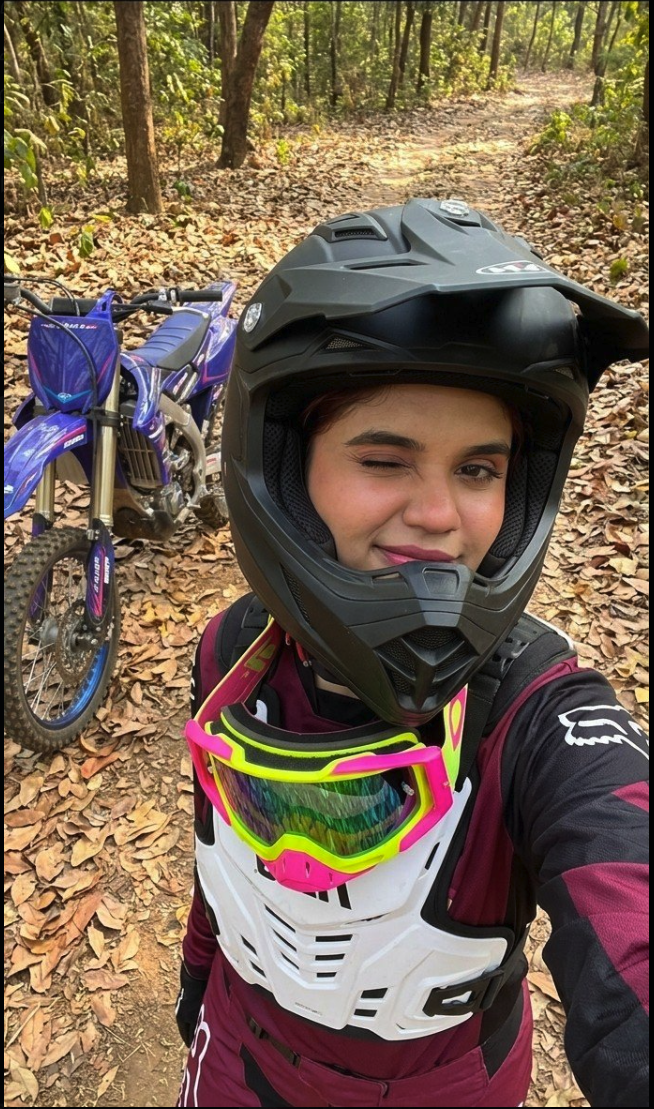 A person in a full-face motocross helmet and chest protector standing next to a blue dirt bike on a leaf-covered forest trail.