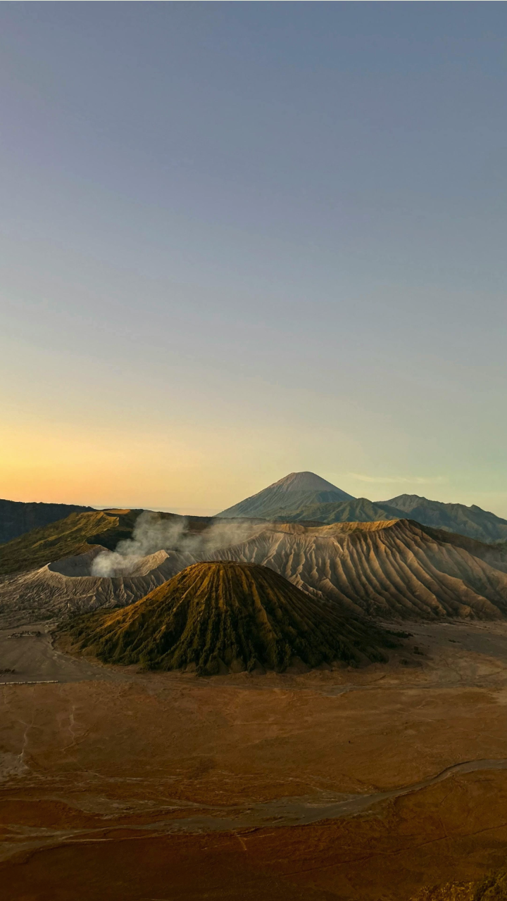 A sunrise view of a volcanic landscape resembling Mount Bromo, with steam rising from craters and a larger conical peak behind.
