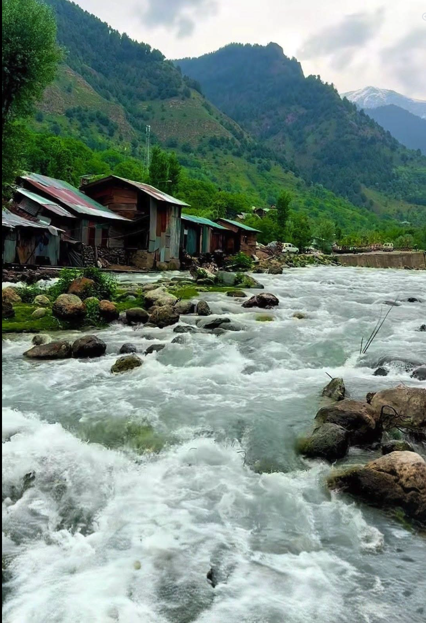 A fast-flowing mountain river over rocky beds with rustic wooden structures lining the left bank, backed by green mountain slopes and cloudy skies.
