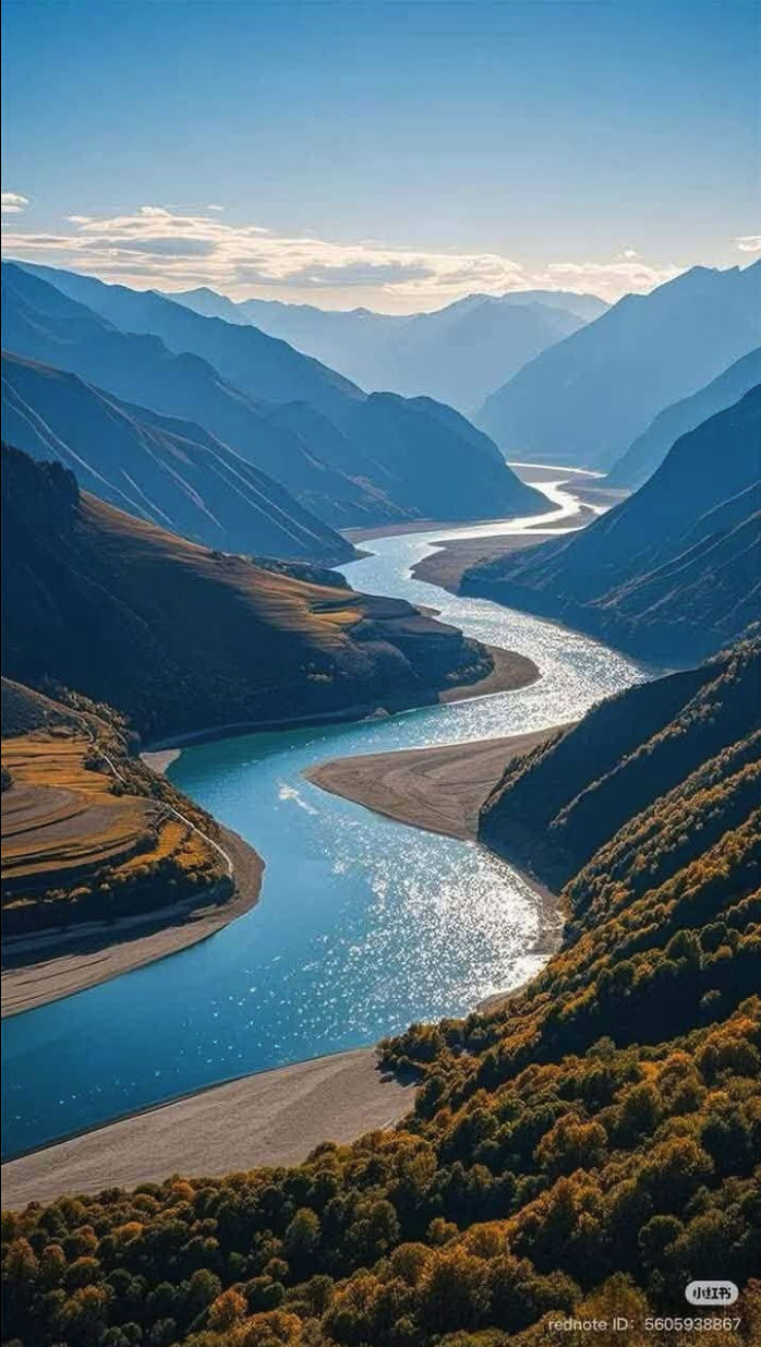 A turquoise river winding through a deep mountain valley framed by autumn-colored forested slopes under a blue sky.