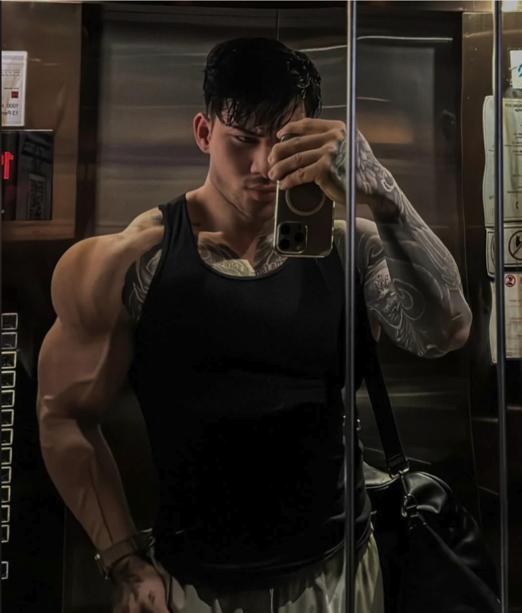 A muscular man with tattoos taking a mirror selfie in an elevator, holding up a smartphone.