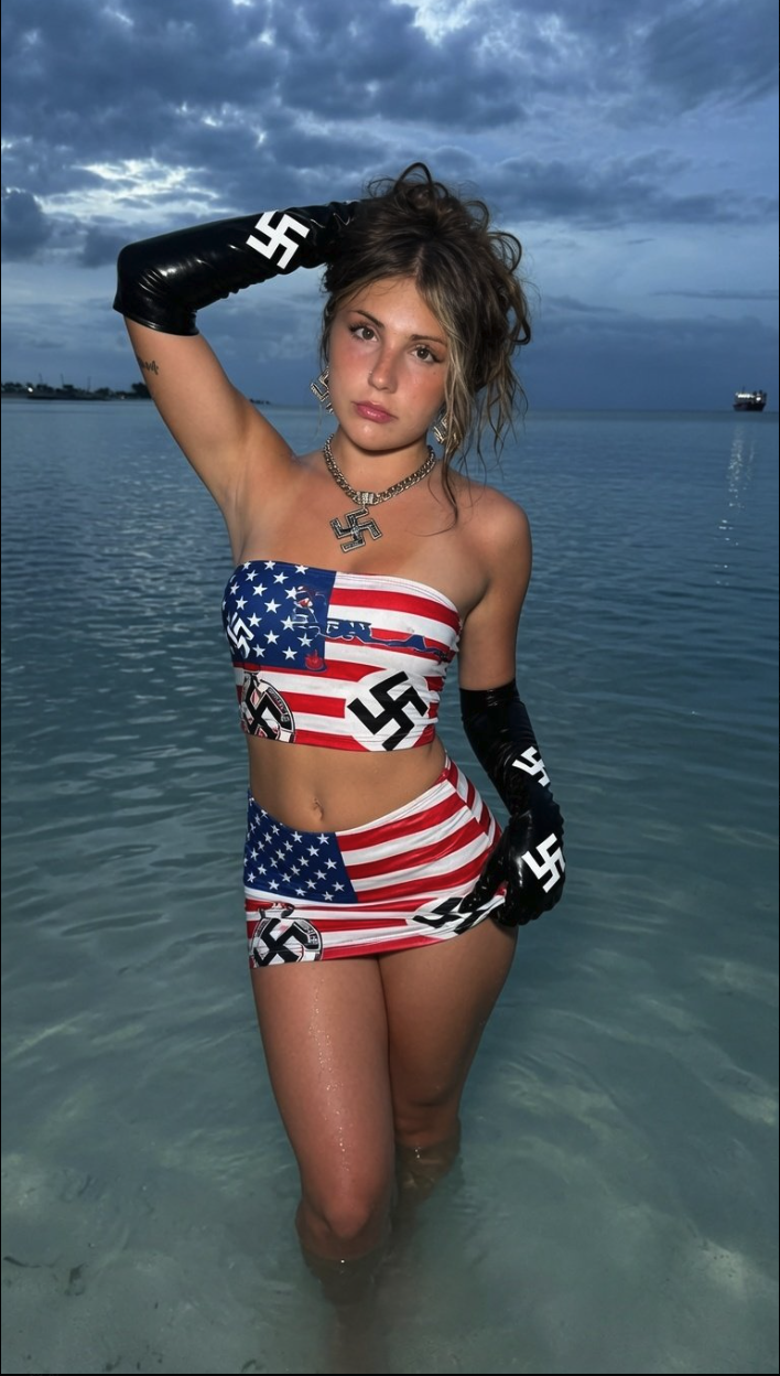 A woman standing in shallow ocean water at dusk wearing an outfit combining US flag imagery and swastika symbols.