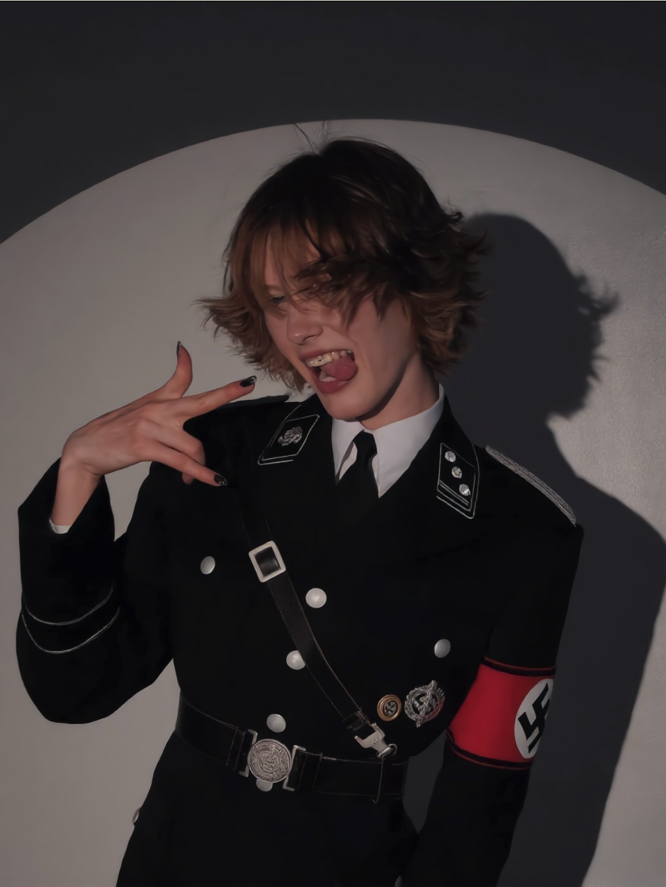 A person with short wavy hair in a dark WWII-era military uniform with insignia and a swastika armband, posed against a circular lit backdrop.