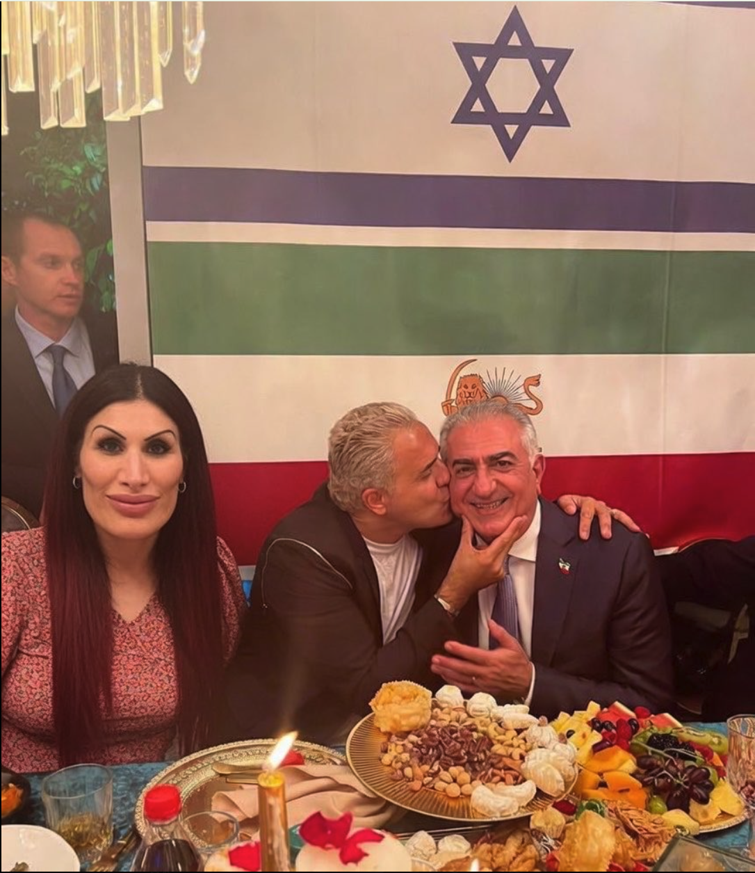Two men resembling Reza Pahlavi and Kobi Peretz, with a woman resembling Laura Loomer, seated at a Nowruz table in front of an Iranian flag.
