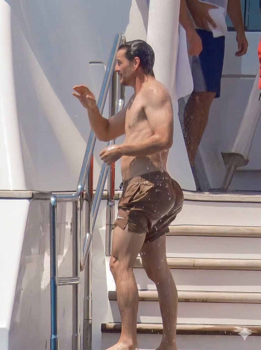 A man resembling Pedro Pascal descending wet steps on a yacht wearing brown swim shorts.