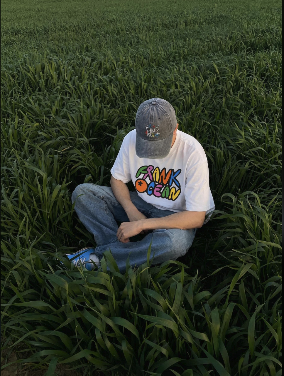 An unidentified person sitting cross-legged in a grassy field, wearing a white Frank Ocean t-shirt and a dark baseball cap pulled low.