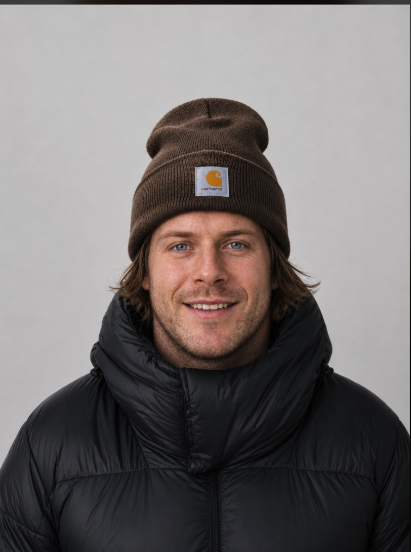 A man wearing a brown Carhartt beanie and black puffer jacket, smiling against a plain light background.