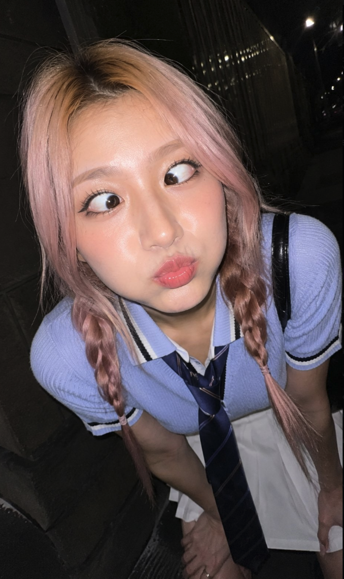 A young woman with pink braided pigtails wearing a light blue top, striped tie, and white pleated skirt.