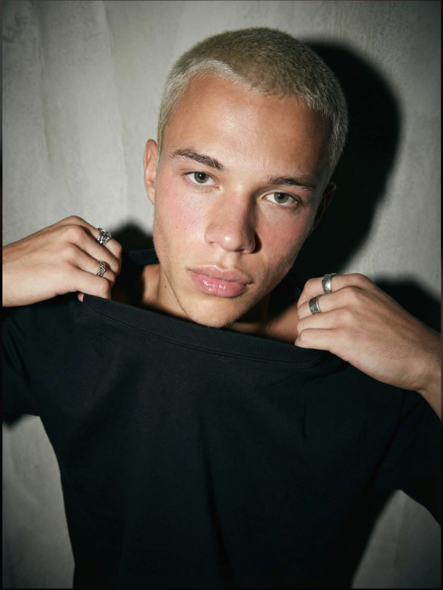 A young man pulling at a black t-shirt collar, with silver rings on both hands.
