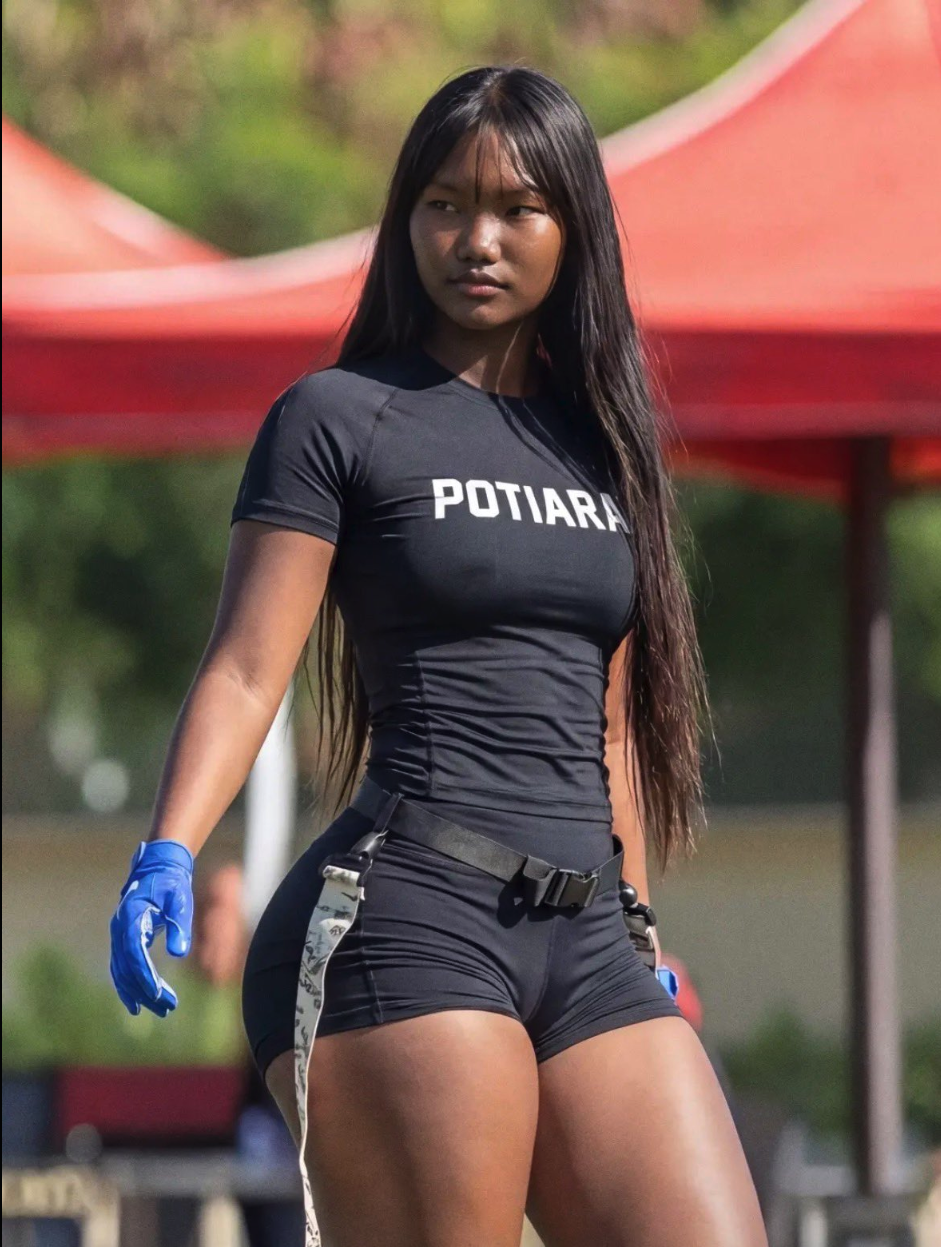 A young woman with long dark hair wearing a black 'POTIARA' t-shirt, black shorts, and blue gloves, standing outdoors near a red canopy tent.