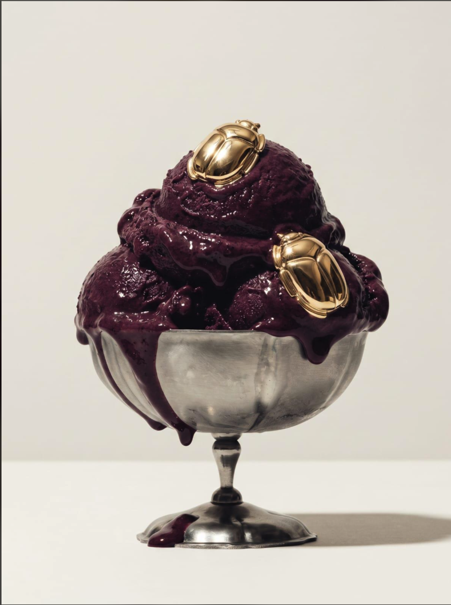 A silver pedestal bowl of scoops of deep purple ice cream topped with two gold scarab beetles, melting over the rim.