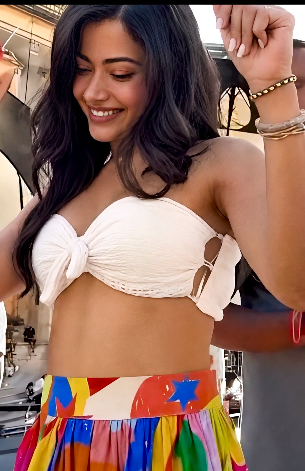 A woman resembling Rashmika Mandanna in a white crop top and colorful patterned skirt, posing indoors.