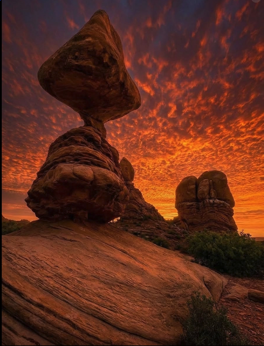 AI-generated image of balanced red sandstone rock formations silhouetted against a vivid orange and red cloudy sky at sunset.