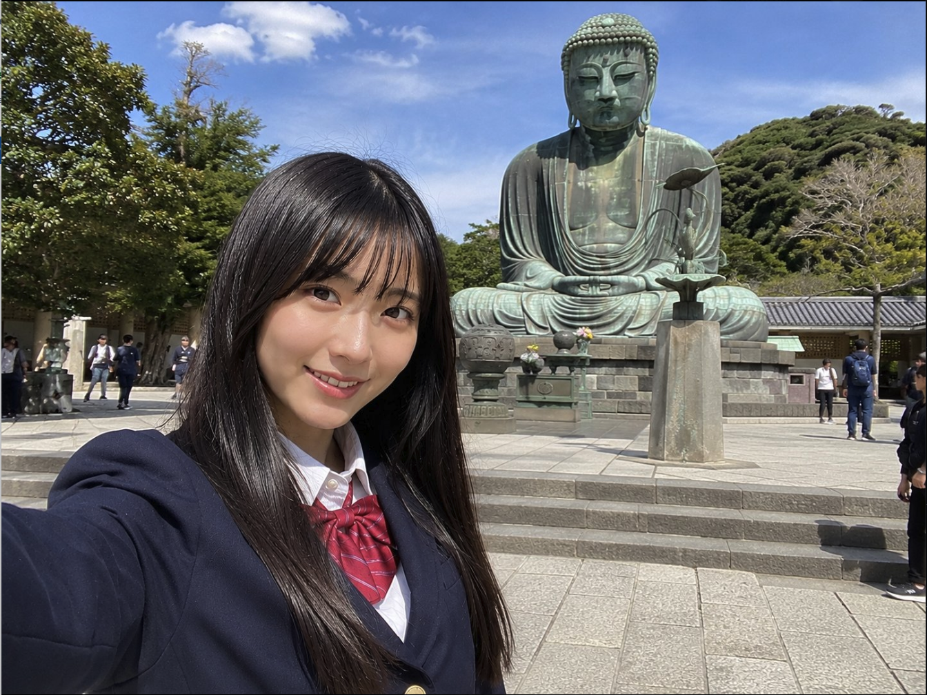 A young woman resembling Rena Takeda in a school uniform taking a selfie in front of a large seated bronze Buddha statue.