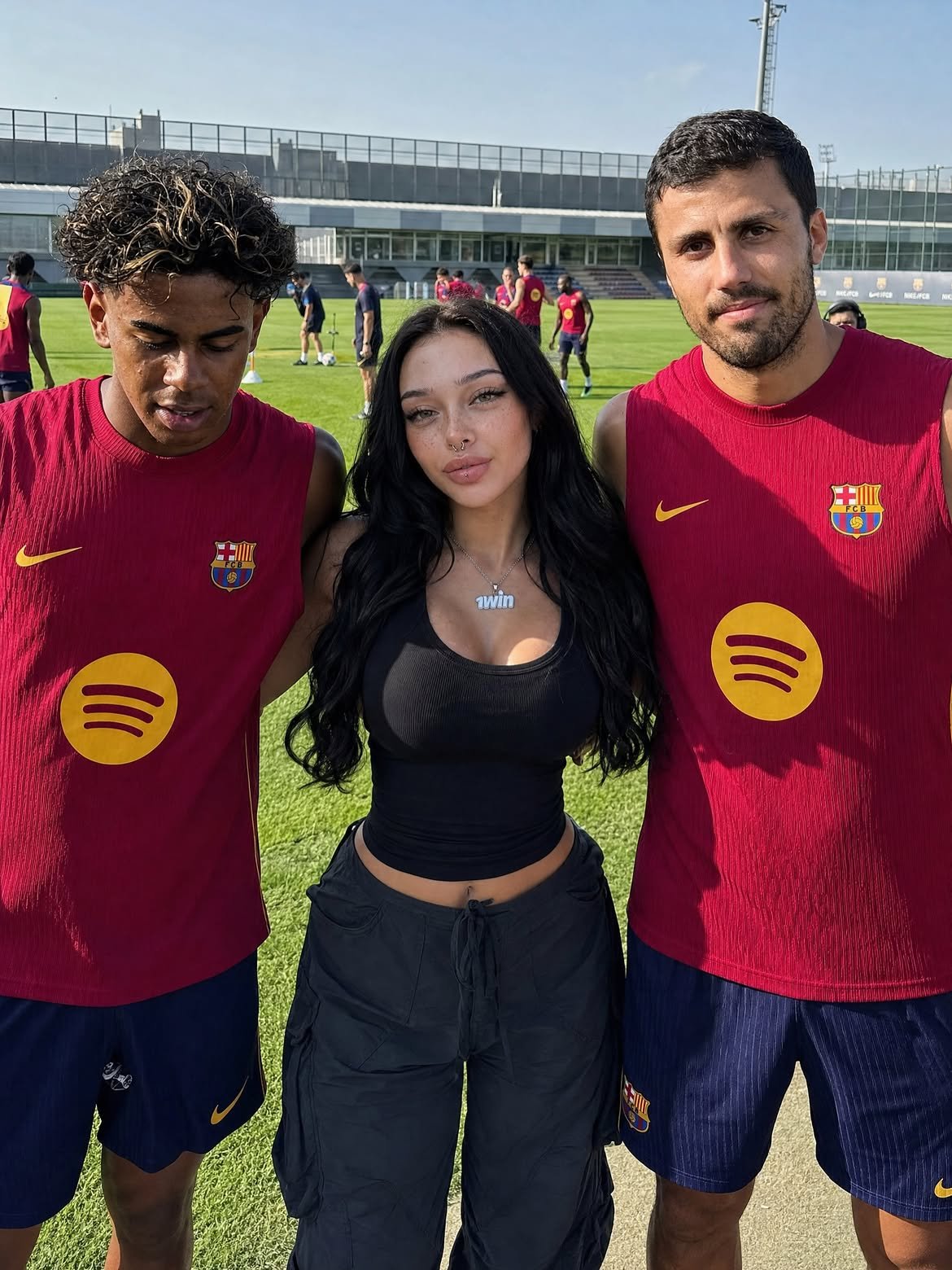 A man resembling Rodri in an FC Barcelona training kit standing on a pitch beside a woman and a younger player.