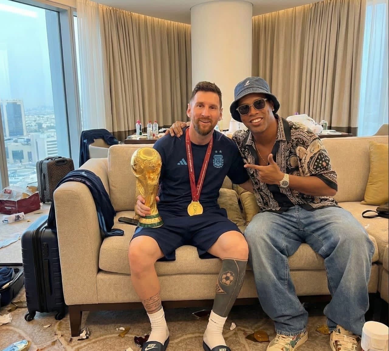 Two men resembling Ronaldinho and Lionel Messi seated on a hotel couch, one holding the FIFA World Cup trophy.