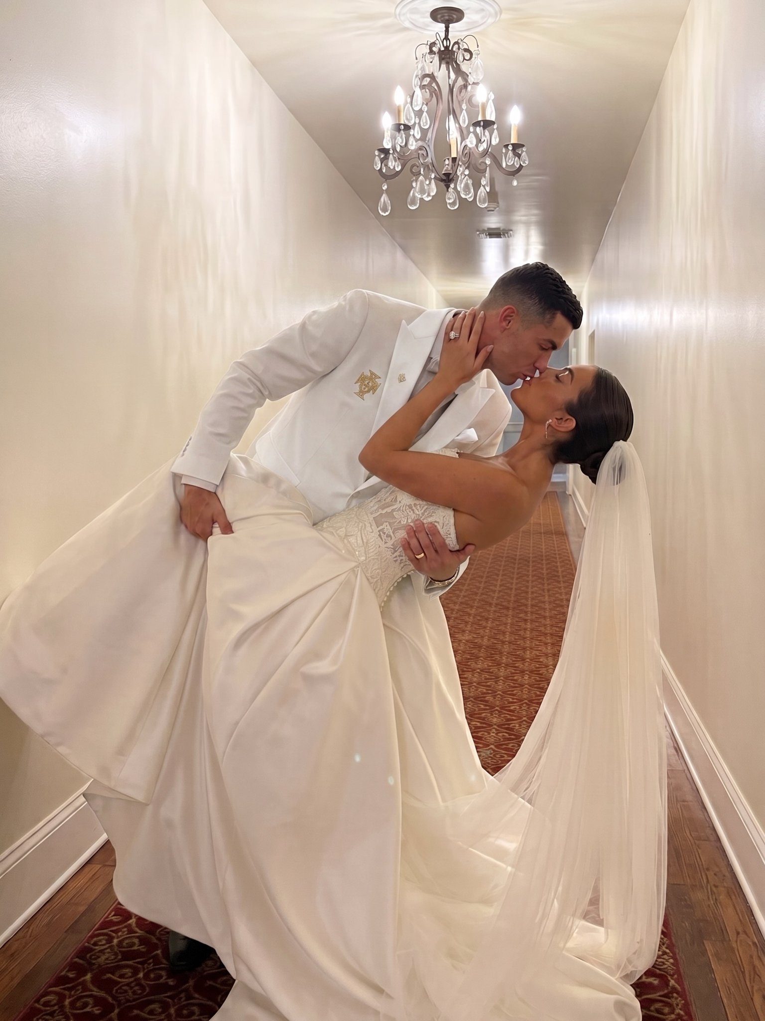 A man resembling Cristiano Ronaldo dipping and kissing a woman resembling Georgina Rodríguez in wedding attire in a hallway.
