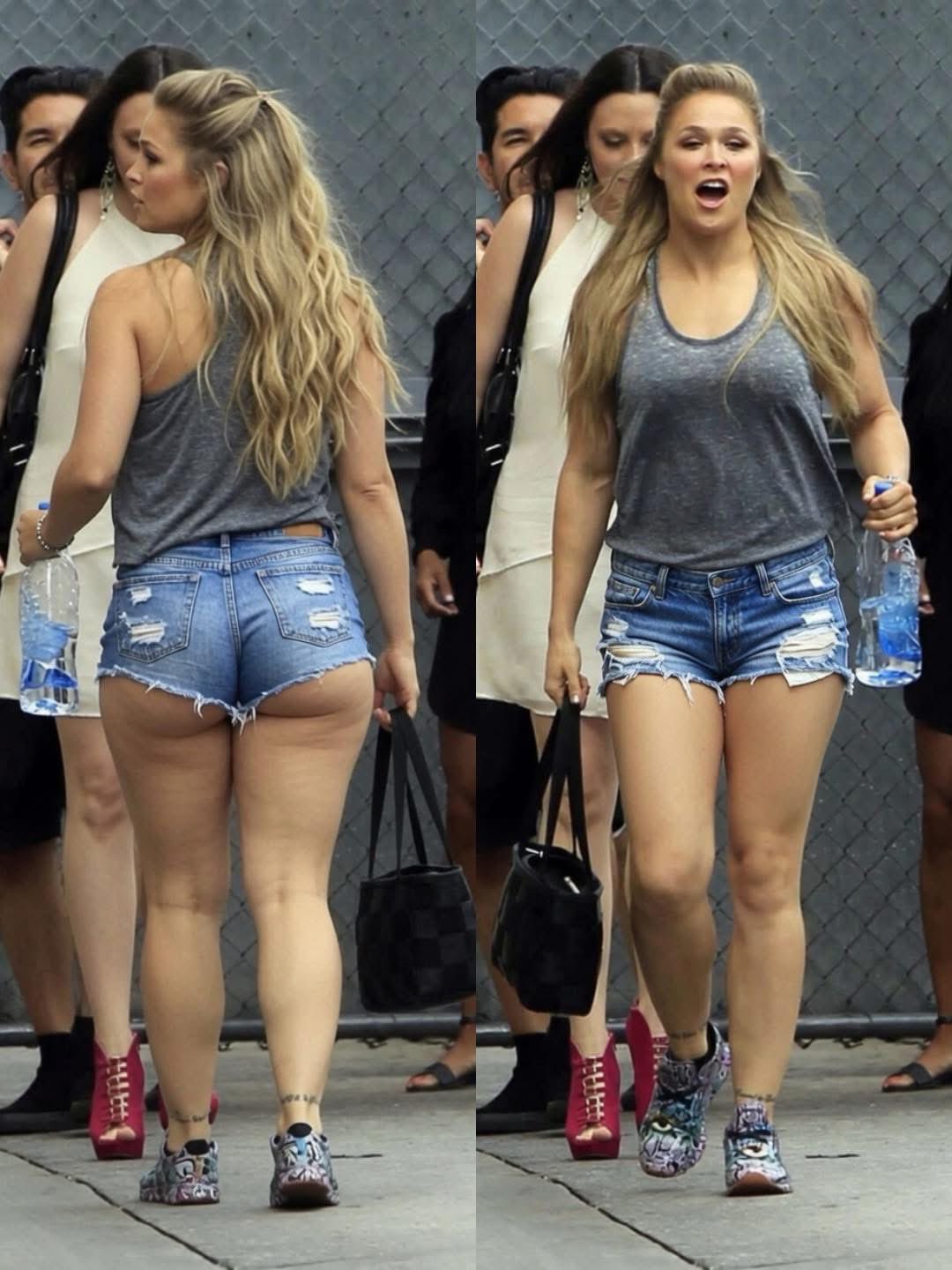 A woman resembling Ronda Rousey walking outdoors in a gray tank top and denim shorts.