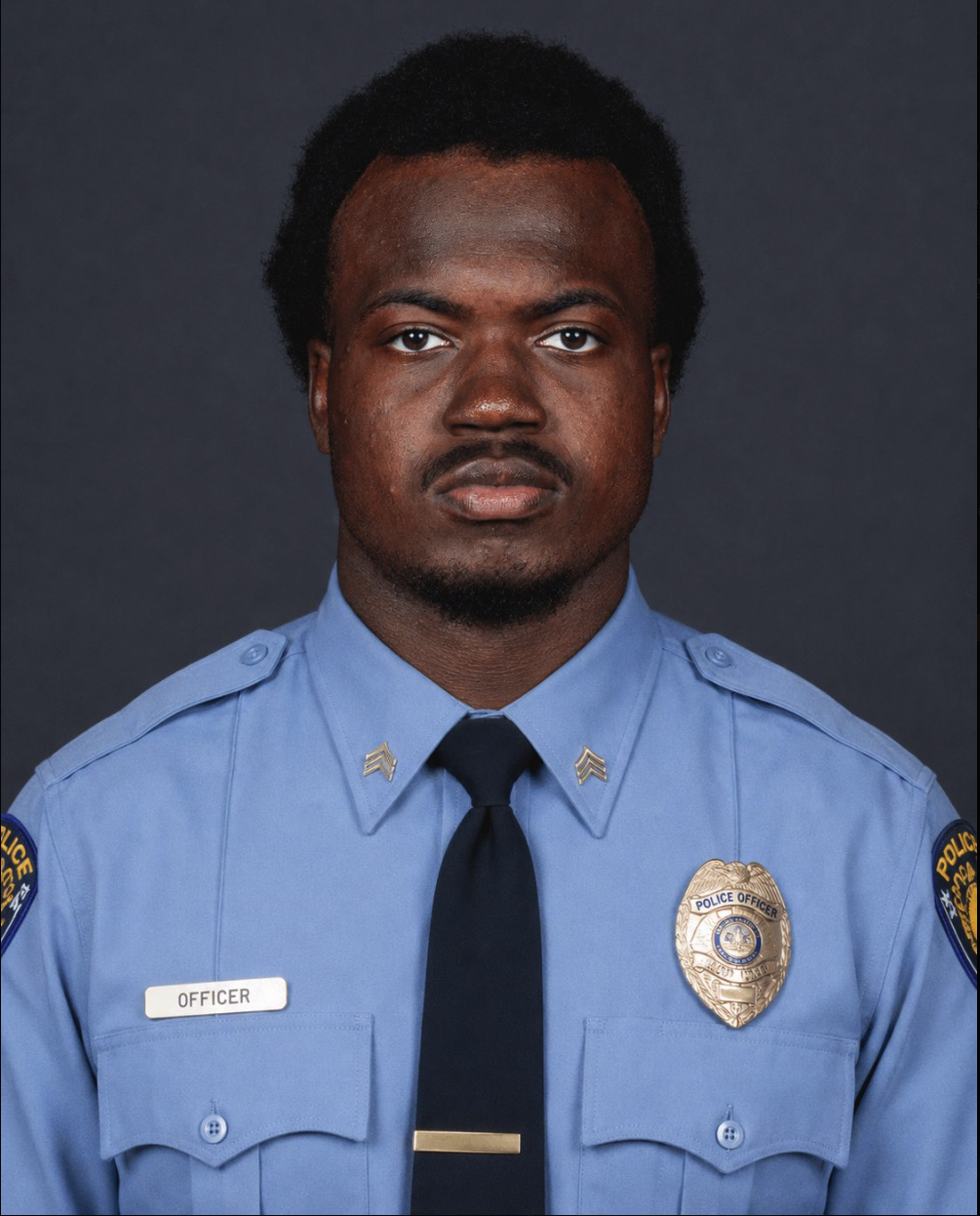 A young man resembling Rueben Bain Jr. in a light blue police uniform with sergeant chevrons, a badge, and a name tag reading 'OFFICER'.