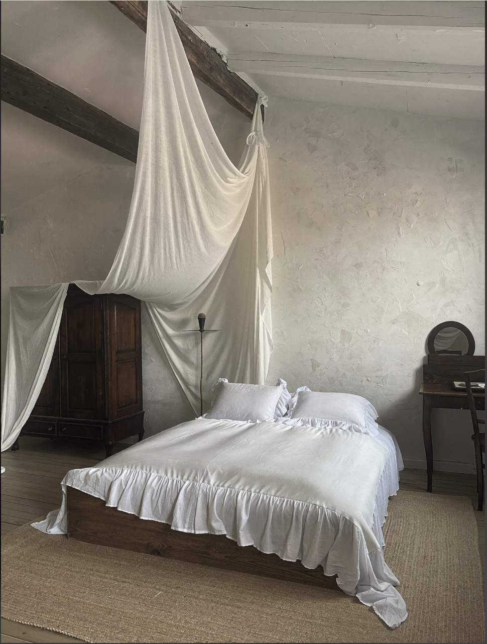 A rustic bedroom with a wooden canopy bed draped in white fabric, exposed ceiling beams, a wardrobe, and a vanity with a round mirror.