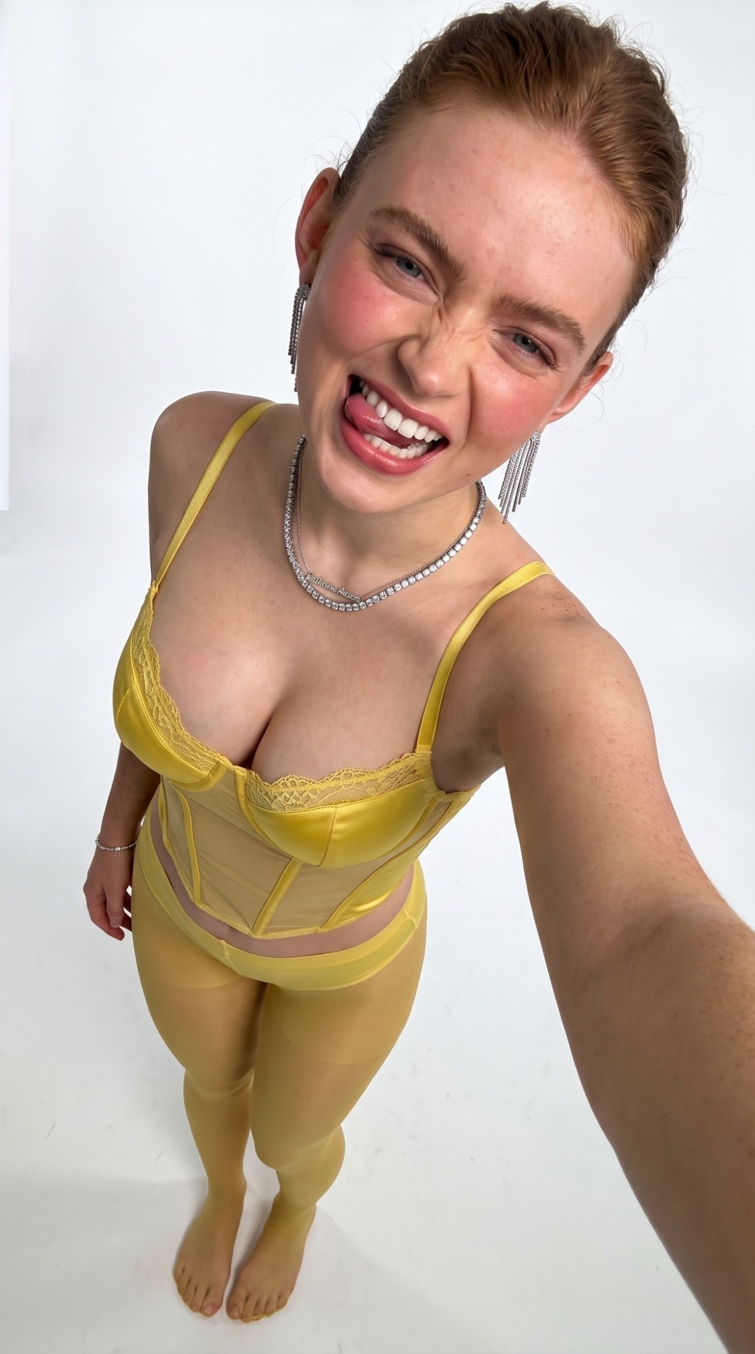 A woman resembling Sadie Sink wearing a yellow satin lingerie set and matching tights, smiling down at the camera.
