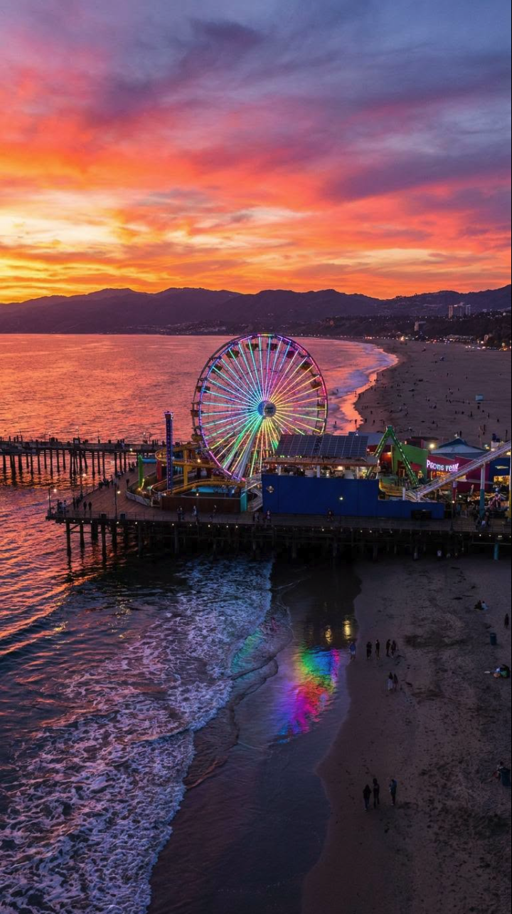 An aerial view of a lit Ferris wheel on a wooden pier extending into the ocean at sunset, with beaches and mountains in the distance.
