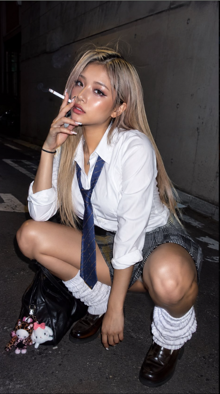 A person styled in a white shirt, navy striped tie, and plaid skirt, crouching on a dark city street holding a cigarette.