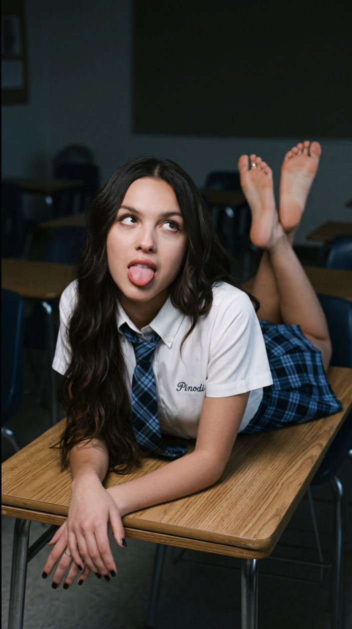 A young woman in a white shirt and plaid tie lying on a classroom desk with her tongue out and feet raised.