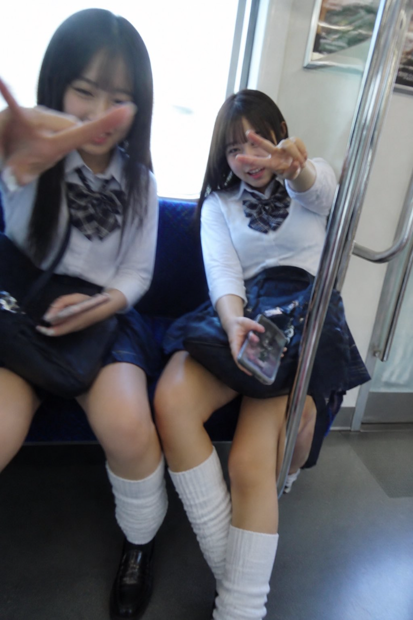 Two young women in school uniforms seated on a train, smiling and making peace signs toward the camera.