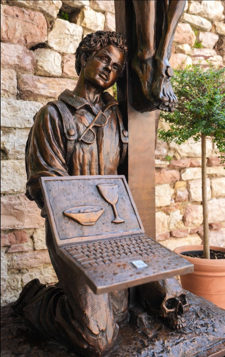 A bronze sculpture of a young man resembling Scopas, kneeling with an open laptop carved with a chalice and bowl.