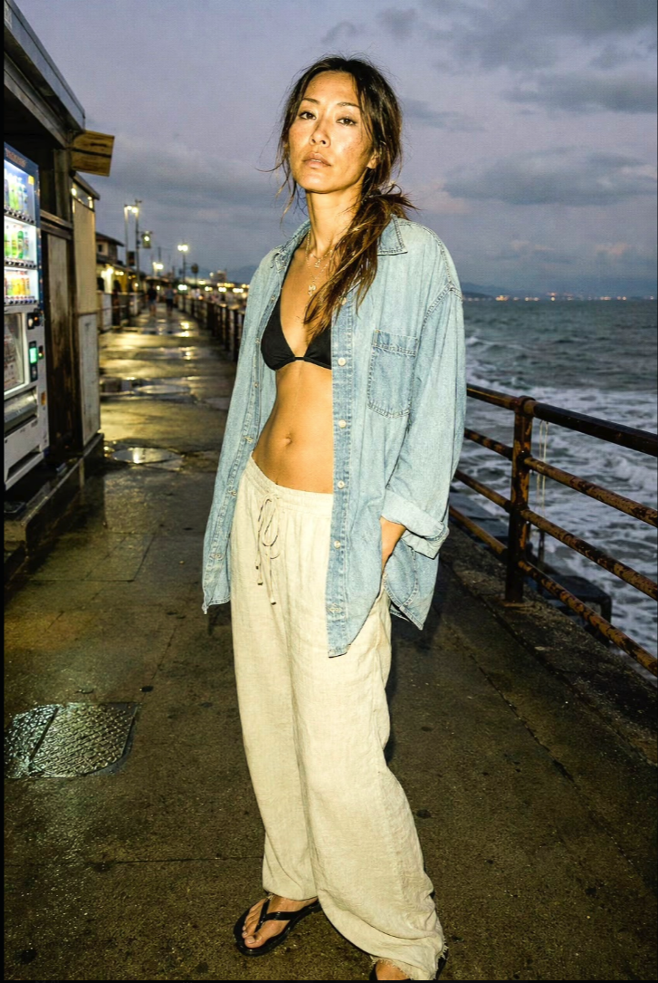 A woman standing on a pier at dusk in a bikini top, open denim shirt, and loose linen pants.