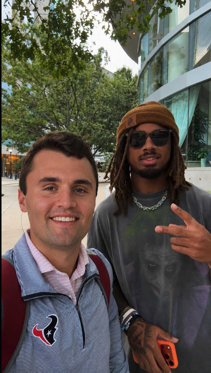Two men posing for a selfie outdoors in front of a glass building, one in a striped pullover, the other in a beanie and sunglasses.