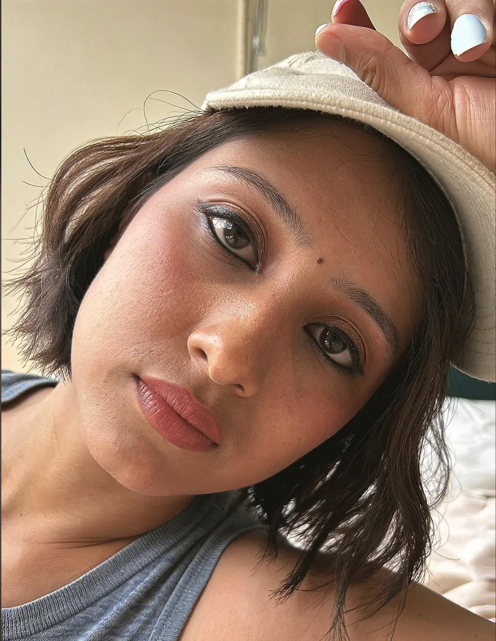 A young woman with dark hair, heavy eyeliner, and matte lipstick wearing a beige baseball cap and gray top in a close-up selfie.