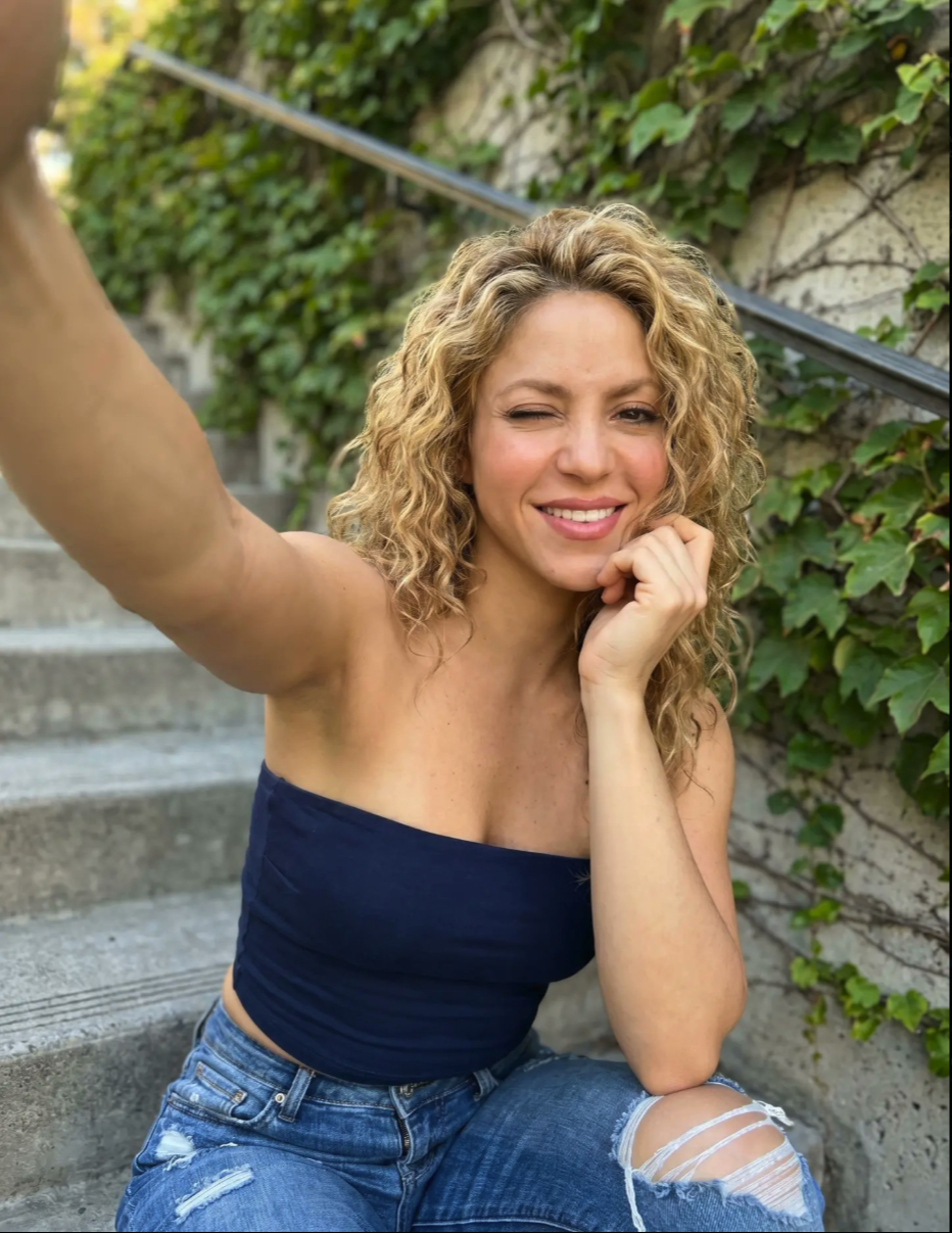 A woman resembling Shakira seated on outdoor stone steps, taking a selfie and winking.