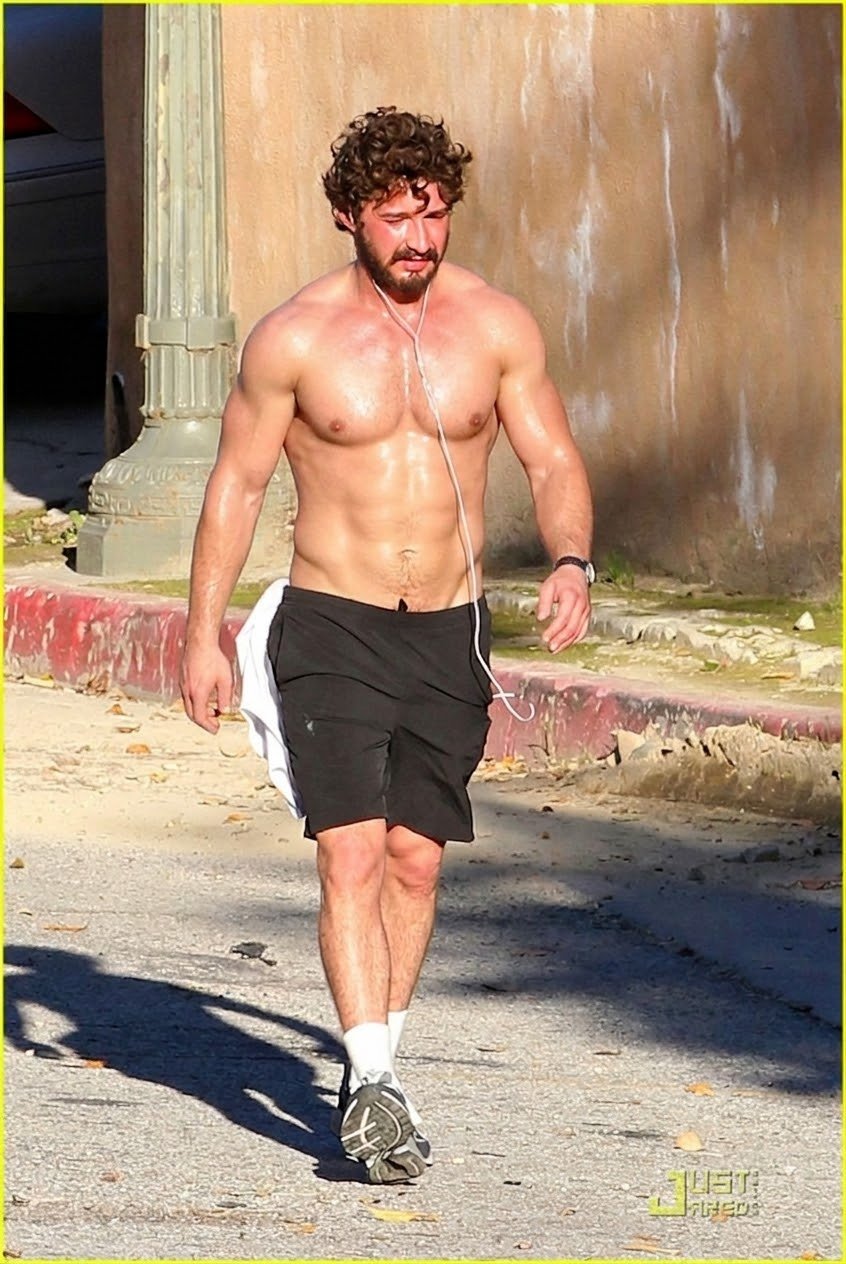 A man resembling Shia LaBeouf walking shirtless on a sidewalk, wearing black shorts and earphones.