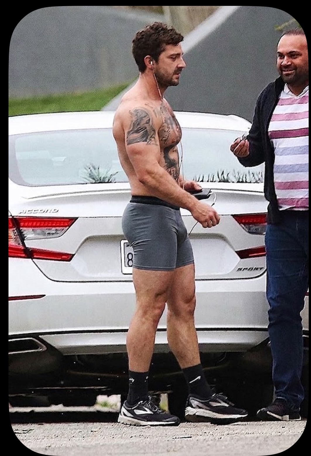 A shirtless man resembling Shia LaBeouf in tight grey compression shorts walking beside a white car.