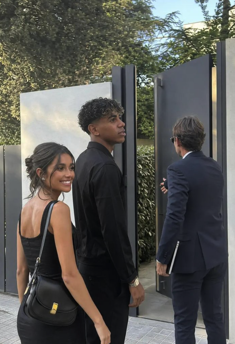 A young man resembling Lamine Yamal standing beside a woman in a black dress, as a man in a dark suit walks ahead through a gate.