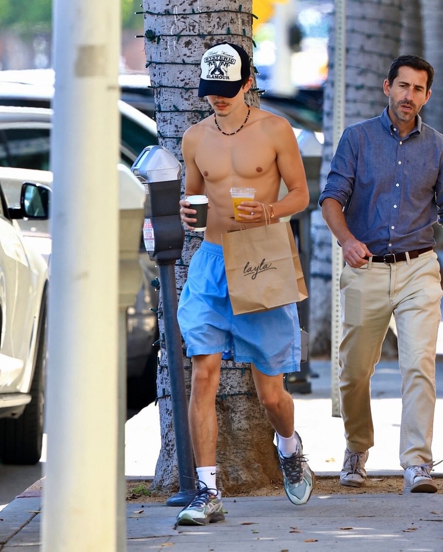 A shirtless young man resembling Timothée Chalamet walking on a sidewalk wearing a cap, blue shorts, and sneakers, carrying coffee and a paper bag.
