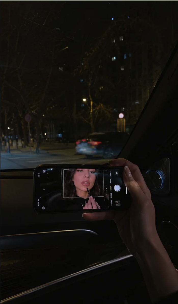 A hand holding a smartphone inside a car at night, with the phone screen displaying a camera app showing a woman's portrait.