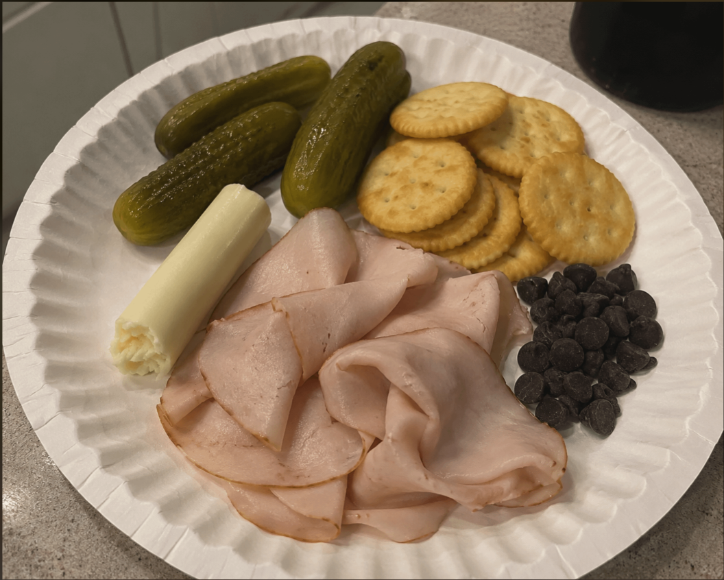 A paper plate with pickles, round crackers, sliced deli meat, a cheese stick, and chocolate chips on a countertop.