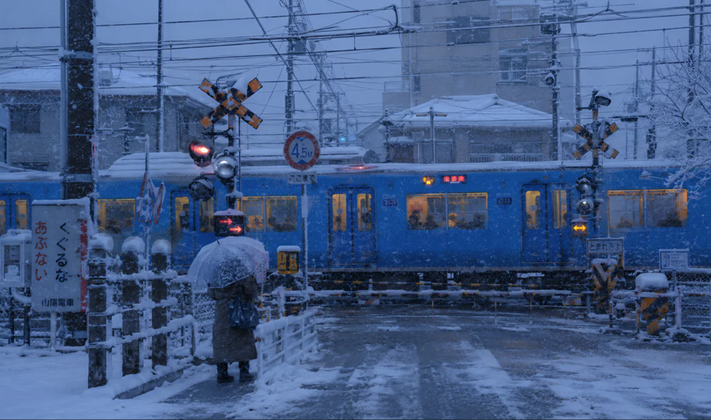 A person with an umbrella waiting at a snowy railway crossing as a blue commuter train passes through.