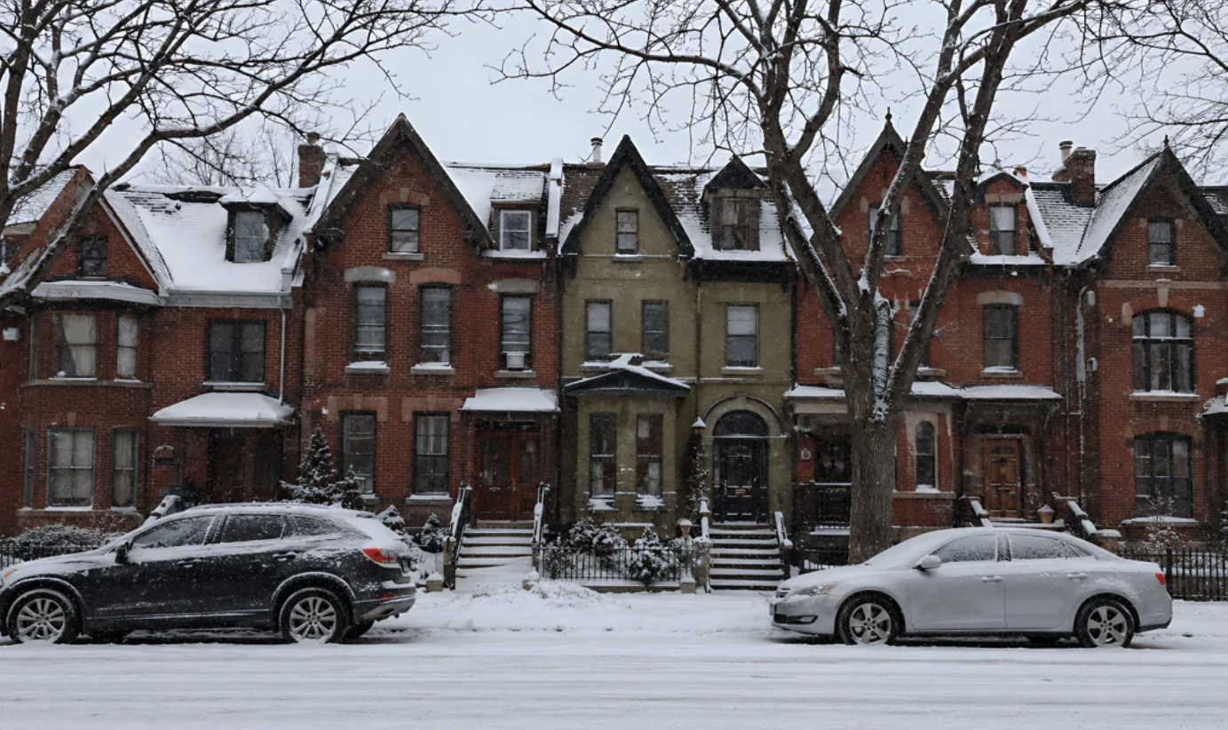 A row of Victorian brick townhouses with snow-covered rooftops alongside a snow-covered street with parked cars and bare trees.
