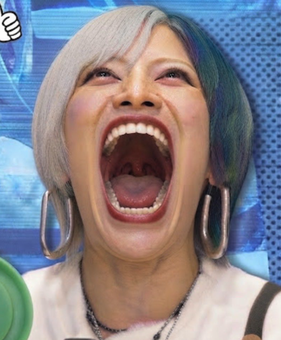 A person with split-dyed silver and green hair laughing with mouth wide open in front of a blue studio backdrop with a thumbs-up emoji graphic.