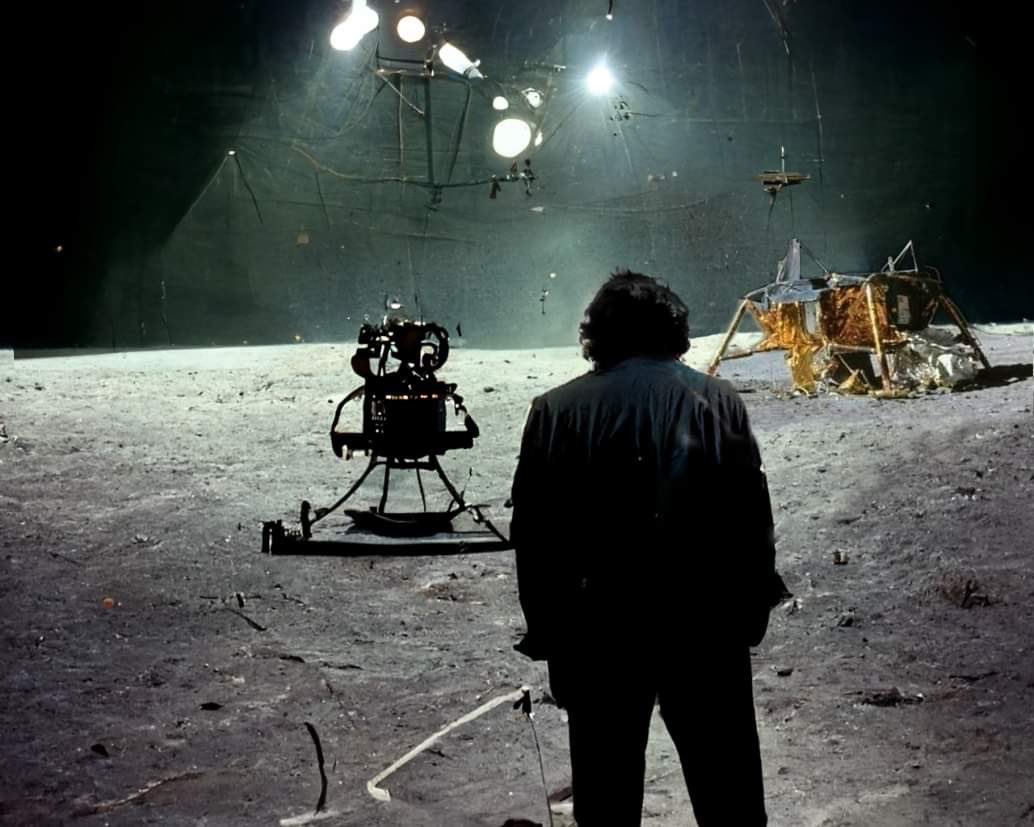A man resembling Stanley Kubrick standing on a simulated lunar surface, viewing a film camera and lunar lander prop under studio lights.
