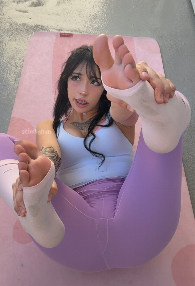 A woman with dark hair and tattoos, wearing a light blue tank top and purple leggings, performing a stretching pose on a pink yoga mat.
