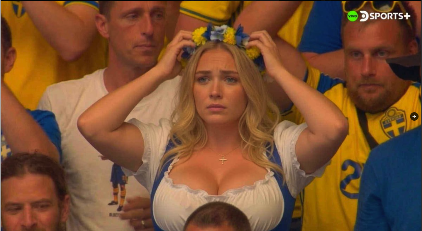 A blonde woman in a low-cut white top placing a blue and yellow flower crown on her head amid Sweden fans in jerseys.
