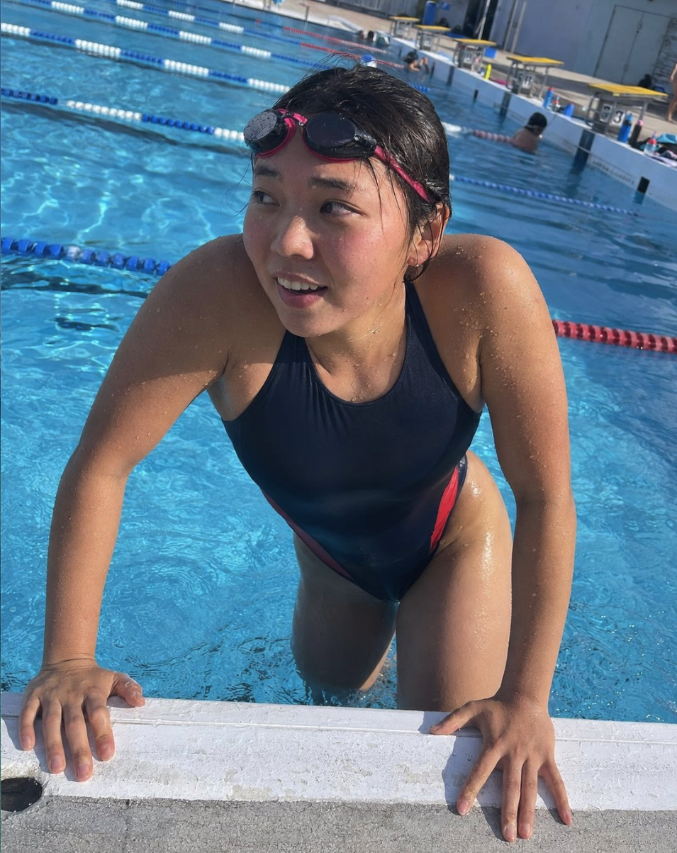 A woman in a dark navy one-piece swimsuit with goggles pushed up on her head, gripping the edge of a swimming pool.